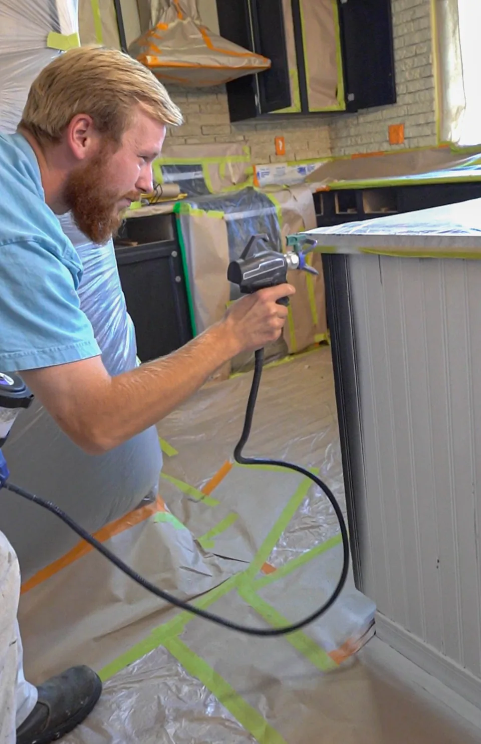 Painter spraying a white panelled kitchen island, with the floor and counters covered in plastic and tape