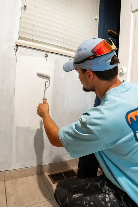 Painter rolling white paint on an interior wall under a window, kneeling on a tiled floor