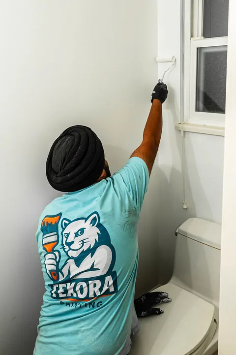 Painter in a Texora T-shirt and black gloves rolling a bathroom wall next to a window